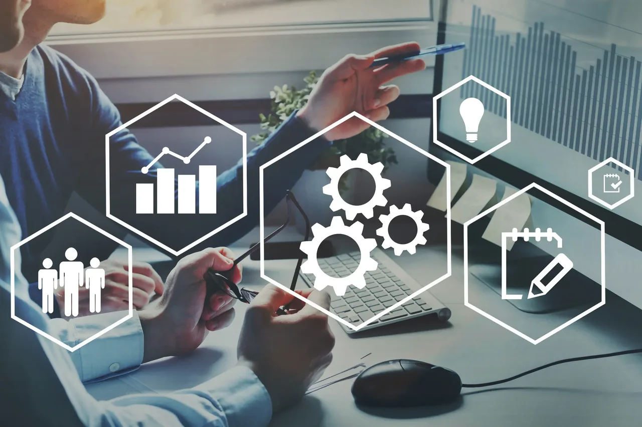 Two colleagues reviewing plans at a desk with hexagonal icons for people, analytics and process gears overlaid on the scene