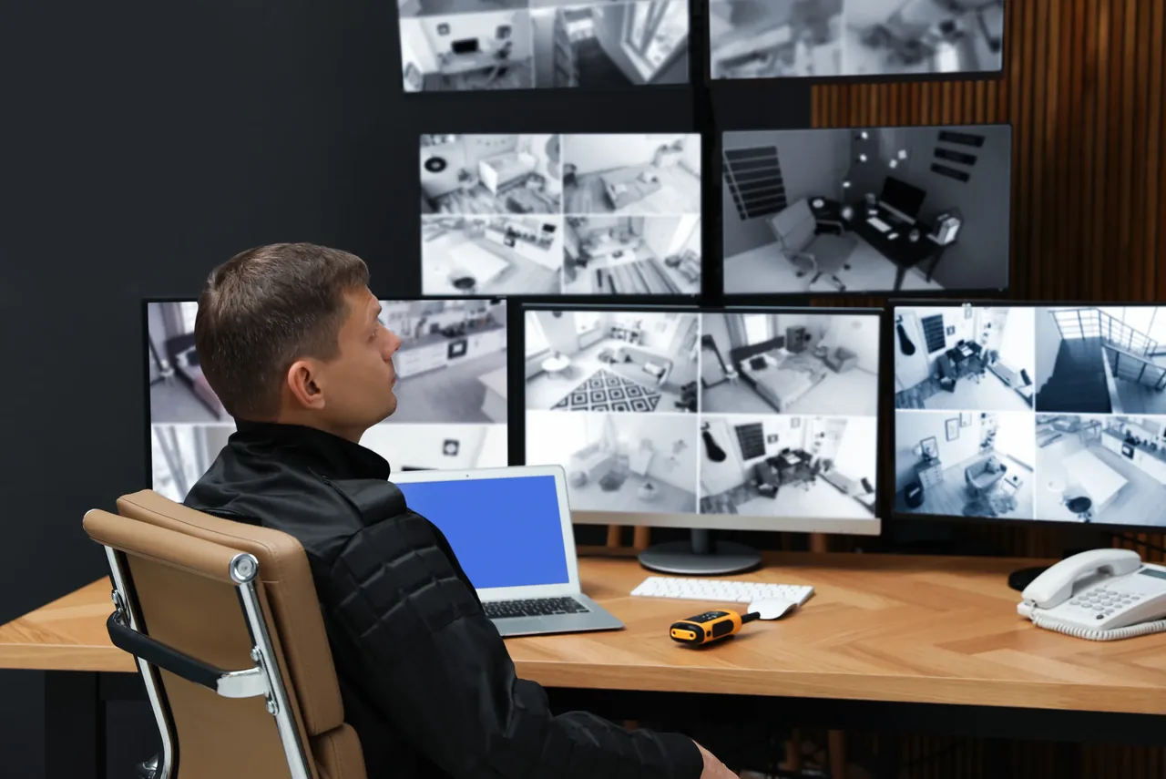 Security operator seated at a control-room desk in front of a wall of monitors displaying multiple live CCTV camera feeds