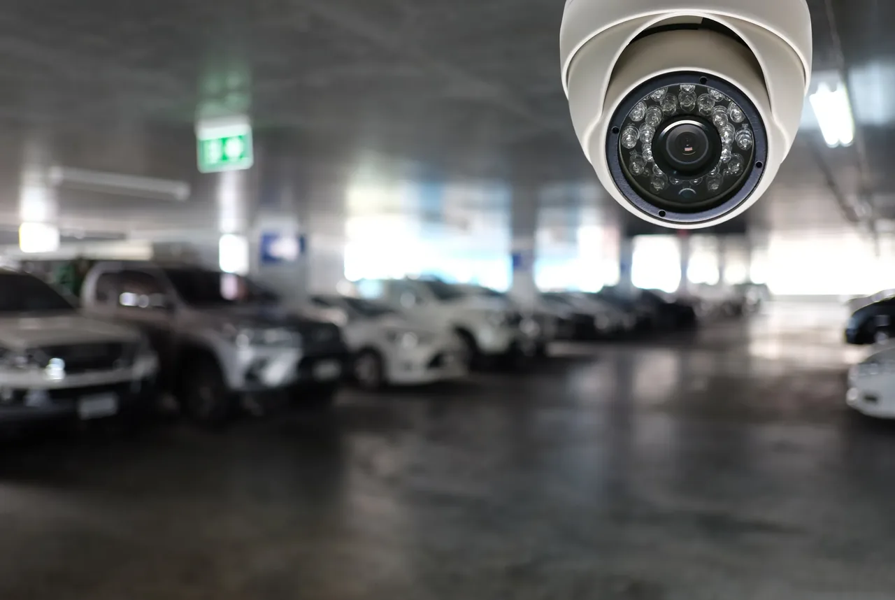 White dome surveillance camera mounted in a covered car park with rows of parked vehicles behind it
